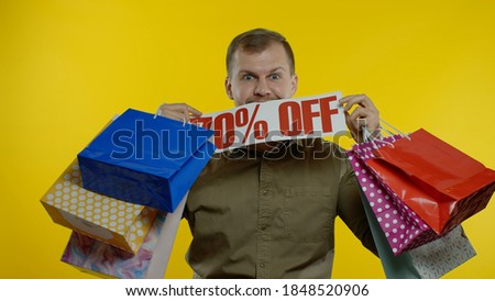Joyful man showing Up To 70 percent Off inscription sign and shopping bags, smiling, looking satisfied with low prices, shopping on big sale day. Yellow background. Black Friday concept
