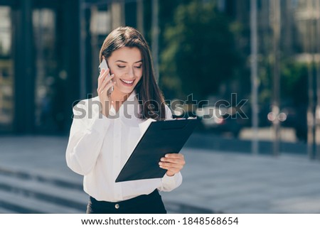 Photo of smiling nice young woman wear white formal shirt holding clipboard talking modern gadget outdoors