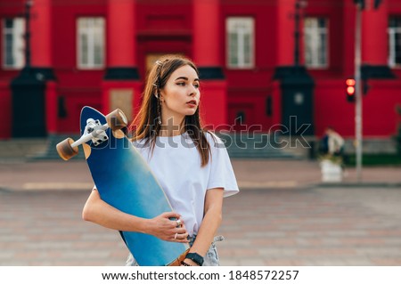 Stylish female skater in casual clothes posing at camera with a longboard in her hands on a background of bright architecture, looking to the side. Skateboarding