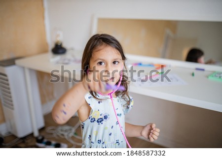 Picture a little brunette girl playing with a pink toy stethoscope and pointing a purple toy thermometer towards the camera, with a blurry mirror and a white desk with coloring images behind her