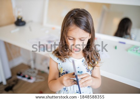 Picture of a little brunette girl looking at a closed American passport in her hands, and with a mirror and a white desk full of coloring images and color pencils behind her