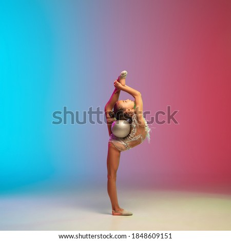 Dynamic. Little caucasian girl, rhytmic gymnast training, performing isolated on gradient blue-red studio background in neon. Graceful and flexible, strong child. Concept of sport, motion, action.