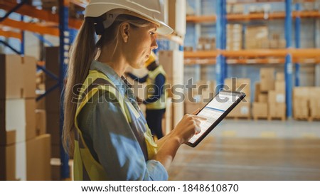 Professional Female Worker Wearing Hard Hat Uses Digital Tablet Computer with Inventory Checking Software in the Retail Warehouse full of Shelves with Goods. Delivery, Distribution Center. Royalty-Free Stock Photo #1848610870