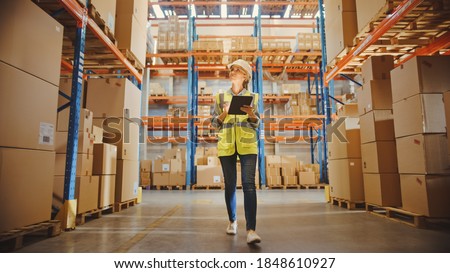 Professional Female Worker Wearing Hard Hat Checks Stock and Inventory with Digital Tablet Computer Walks in the Retail Warehouse full of Shelves with Goods. Working in Delivery, Distribution Center Royalty-Free Stock Photo #1848610927