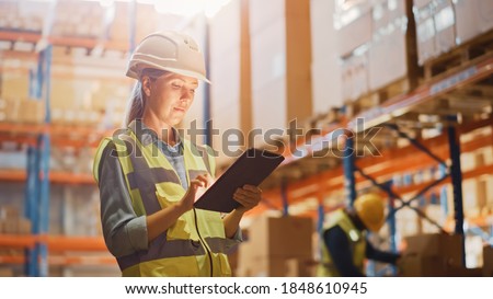 Professional Female Worker Wearing Hard Hat Checks Stock and Inventory with Digital Tablet Computer in the Retail Warehouse full of Shelves with Goods. Working in Logistics, Distribution Center Royalty-Free Stock Photo #1848610945