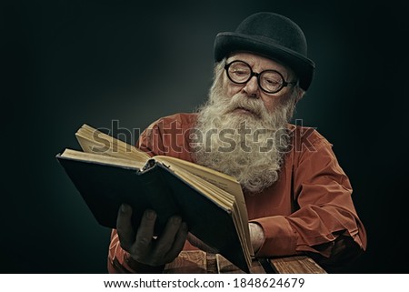 A wise old man with a long gray beard in a bowler hat and glasses is reading an old book. Black background.  Royalty-Free Stock Photo #1848624679