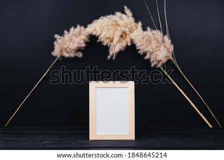 A wooden frame for a poster, inscription, picture, photo stands on a black wooden table. Frame with a clean white sheet. Layout for your design. Frame and reeds around, festive layout.