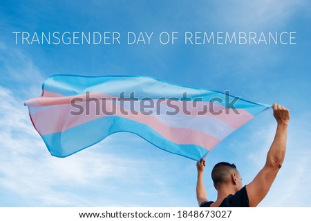 closeup of a young caucasian person, seen from behind, holding a transgender pride flag on the air and the text transgender day of remembrance on the sky Royalty-Free Stock Photo #1848673027