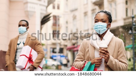 Portrait of pretty happy African American woman in mask walks in snowy town with coffee-to-go after xmas shopping with gifts for new years eve Handsome man with present on background Holidays concept