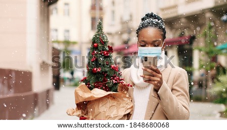 Close up portrait of happy African American young woman in mask standing in snowy decorated city and typing on smartphone. Beautiful female with little new year tree outdoor texting on cellphone