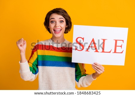 Photo of young cheerful hooray woman stand hold placard sale hand fist wear rainbow sweater isolated yellow color background