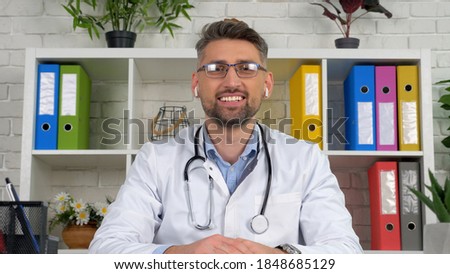 Webcam view smiling man remote doctor wears white medical coat wireless earphones sits on chair at desk in hospital office looks camera consult online distance video call chat laptop computer screen