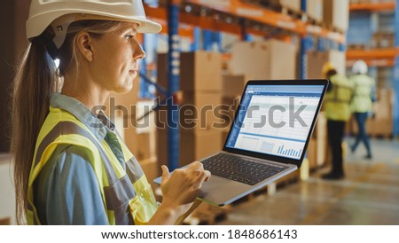 Professional Female Worker Wearing Hard Hat Holds Laptop Computer with Screen Showing Inventory Checking Software in the Retail Warehouse full of Shelves with Goods. Over the Shoulder Side View Royalty-Free Stock Photo #1848686143