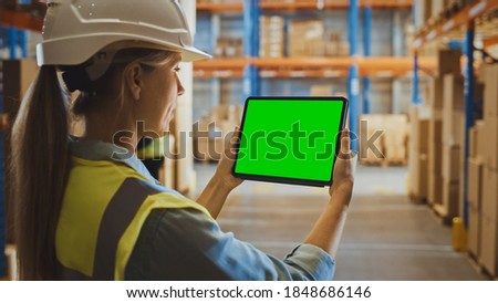 Professional Female Worker Wearing Hard Hat Uses Digital Tablet Computer with Green Chroma Key Screen in Landscape Mode in the Retail Warehouse full of Shelves with Goods. Over the Shoulder view