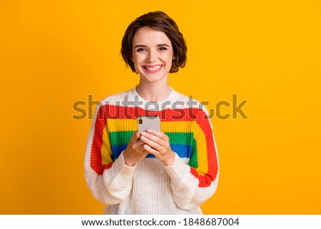 Photo of positive young girl hold telephone beaming smile camera wear striped sweater isolated yellow color background