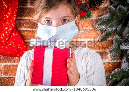 Little Cute Caucasian Blond Girl wearing mask with Red Gift Box with Festive Ribbon, Christmas coronavirus and pandemic concept Home background with fir tree, Gifts, Decorations. copy space
