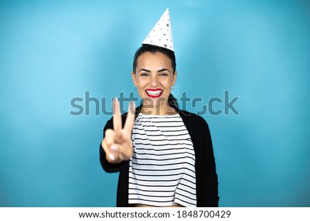 Young beautiful woman wearing a birthday hat over insolated blue background showing and pointing up with fingers number two while smiling confident and happy