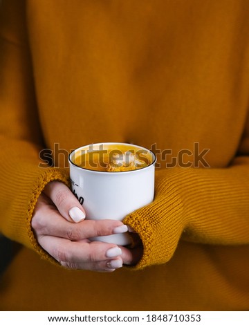 Cup with pumpkin cream soup in girl hands on orange background with copy space for your text