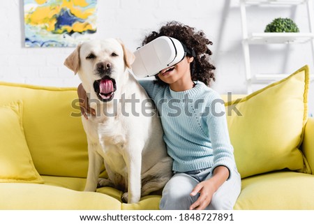 Curly african american girl in vr headset embracing dog yawning, while sitting on sofa on blurred background