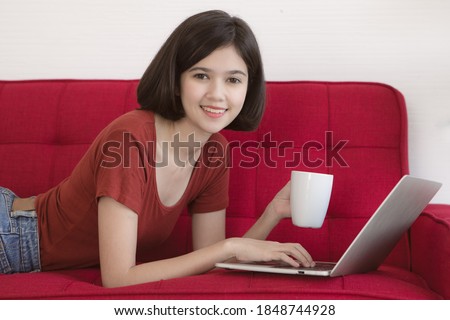 Half race Thai-German cute girl holding white coffee cup laying on red sofa and using laptop notebook computer work from home with happiness feeling.