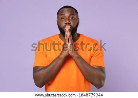 Pleading begging young african american man 20s wearing basic casual orange blank empty t-shirt standing holding hands folded in prayer isolated on pastel violet colour background, studio portrait