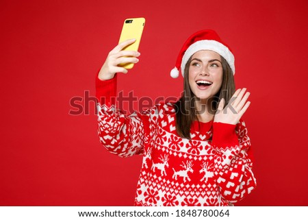 Excited young Santa woman in sweater, Christmas hat doing selfie shot on mobile phone waving greeting with hand isolated on red background studio. Happy New Year celebration merry holiday concept