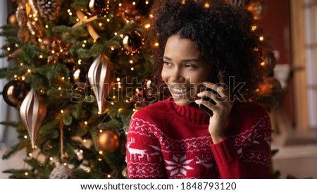 Close up smiling African American young woman talking on phone, sitting near festive Christmas tree at home, happy female holding smartphone, chatting with friends or relatives, congratulating