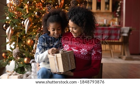 Close up happy African American woman with adorable daughter opening Christmas gift box, sitting on floor near festive tree at home, smiling mother and little girl hugging, unpacking present