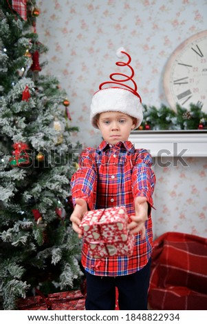 
surprised little boy is holding a red
present box in arms on Christmas tree and clock and fireplace background