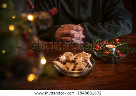 Praying hands of an elderly man on a table with Christmas cookies, candle and festive decoration, lonely holidays during the coronavirus pandemic, copy space, selected focus, narrow depth of field