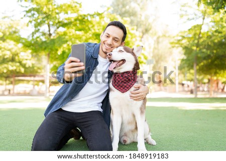 Handsome adult man in his 30s taking a selfie with his cute pet wearing a bandana in the grass at the park