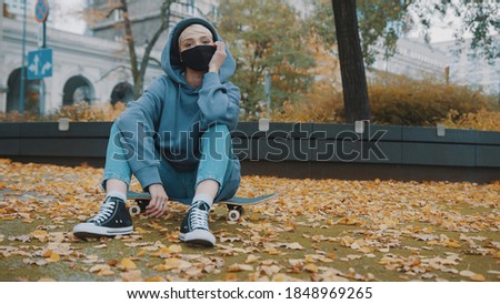 Young hispter woman with face mask and hoodie sitting on the skateboard in the city park in autumn. High quality photo