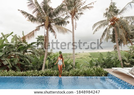 Woman stand on edge luxury infinity pool andenjoying amazing tropical landscape, coconut palm trees and greenery