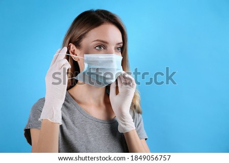 Woman in medical gloves putting on protective face mask against light blue background