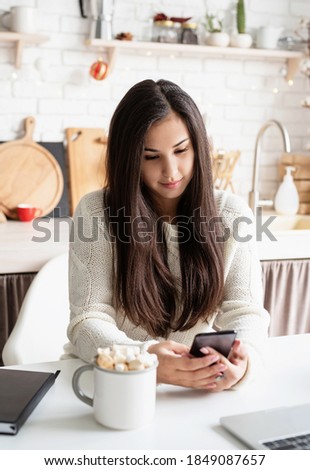 Young brunette woman chatting with friends using her mobile phone at the kitchen