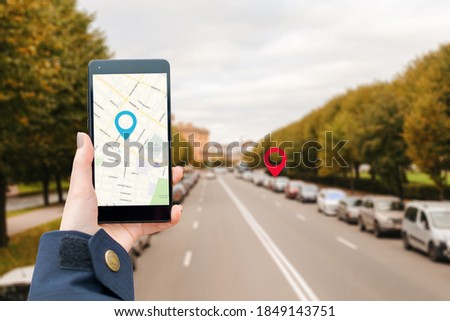 A woman's hand in a jacket holds a smartphone with an online map app. In the background is a blurred road with a cars and icon of location.The concept of Internet navigation and modern technologies