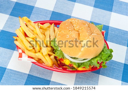 A top down view of a hamburger with fries in a red basket on a picnic tablecloth.