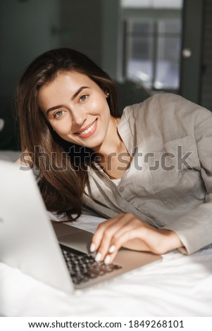 Vertical shot of girl using laptop in bed, smiling at camera and online shopping at home. Young woman relaxing and stream movies.