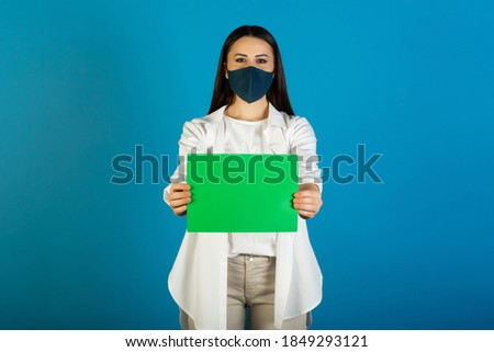 Beautiful girl in medical face mask is hold empty green sheet for your advertisement. Copy space. Coronavirus, COVID-19.