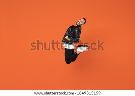 Full length side view of happy joyful young bearded man in basic white t-shirt black leather jacket jumping doing winner gesture clenching fists isolated on orange colour background studio portrait Royalty-Free Stock Photo #1849315159