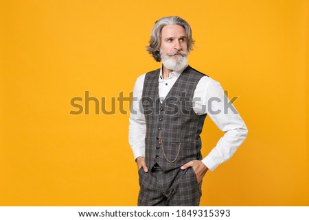 Confident elderly gray-haired bearded business man in checkered suit waistcoat white shirt holding hands in pockets isolated on yellow background studio portrait. Achievement career wealth concept