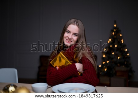 Caucasian girl sitting at a festive table with Christmas candles, hugging a gift box and rejoicing
