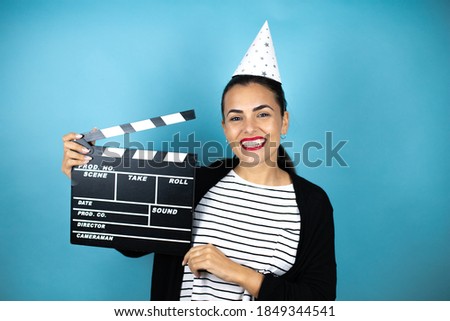 Young beautiful woman wearing a birthday hat over insolated blue background holding clapperboard very happy having fun