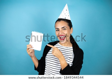Young beautiful woman wearing a birthday hat over insolated blue background smiling and showing blank notebook in her hand