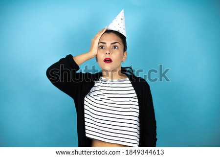 Young beautiful woman wearing a birthday hat over insolated blue background putting one hand on her head smiling like she had forgotten something