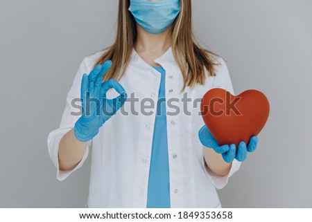 
A female doctor in a white coat, in a blue mask and gloves, holds a red heart in one hand and makes an "okay" gesture with the other.