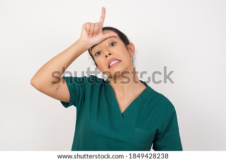 Young arab doctor surgeon woman over isolated white background making fun of people with fingers on forehead doing loser gesture mocking and insulting.