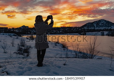 A girl with a camera shoots a colorful evening sunset on a cloudy sky from a height , the silhouette of the photographer in the darkened lighting in the open air with a beautiful view of the glow
