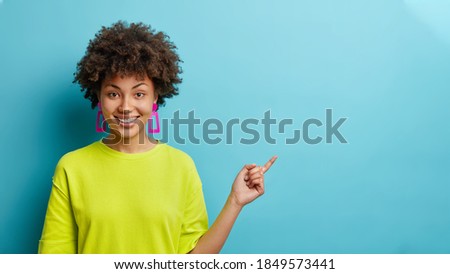 Horizontal shot of cheerful Afro American woman in casual t shirt points away on copy space suggests follow this direction or click on link poses against blue background. Your advertising here