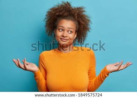 Puzzled young African American woman has curly bushy hair spreads hands in doubt looks aside with unaware face expression dressed in orange long sleeved jumper isolated over blue background.